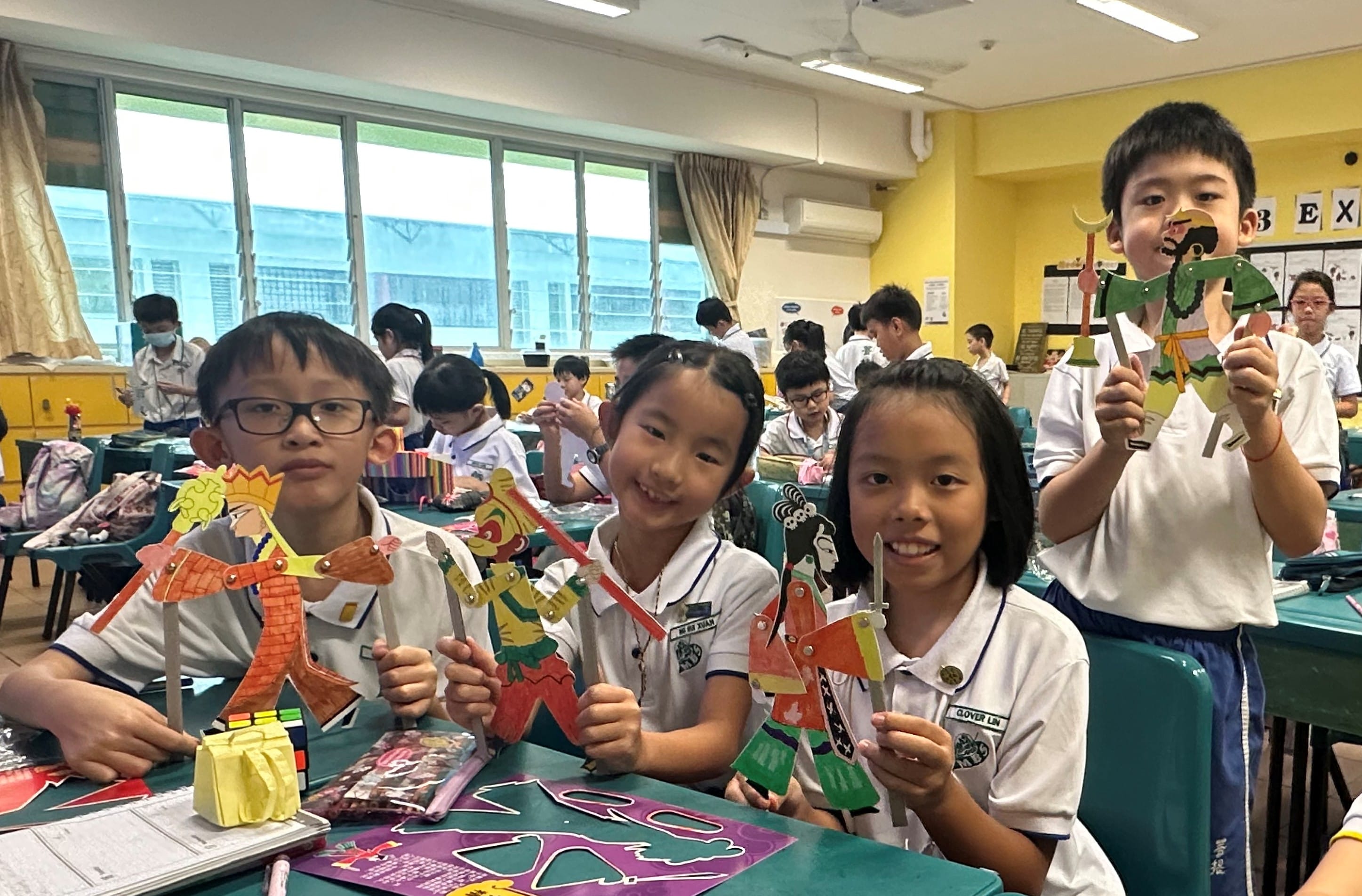 Four smiling children in school uniforms hold up colorful handmade paper puppets in a classroom.